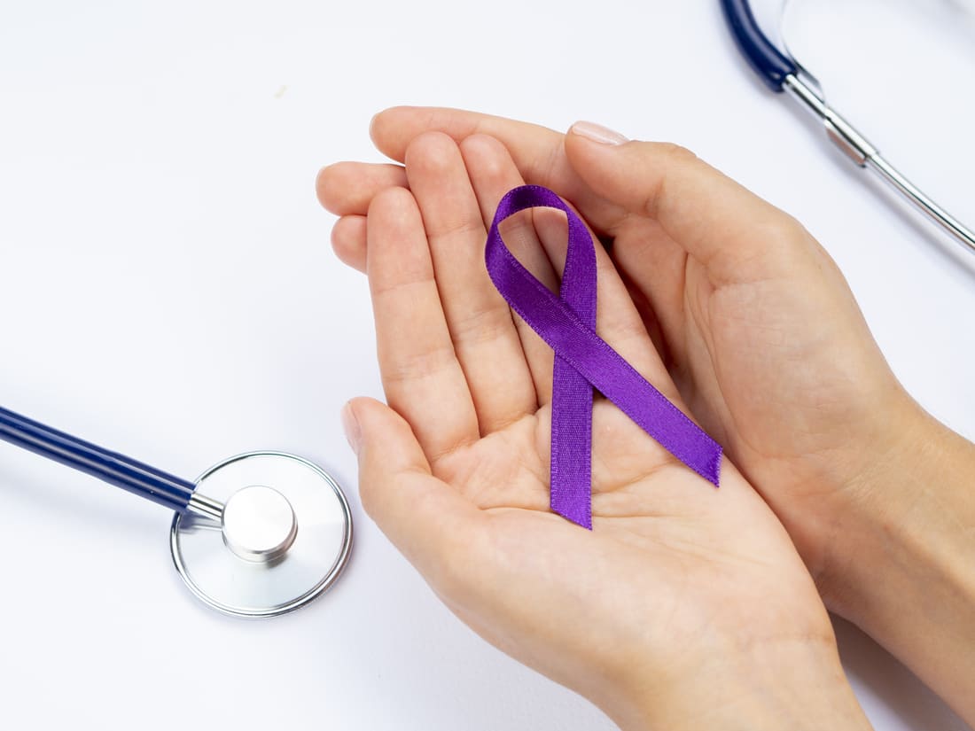 Manos sosteniendo un lazo morado junto a un estetoscopio, tratamientos del cáncer en Colombia